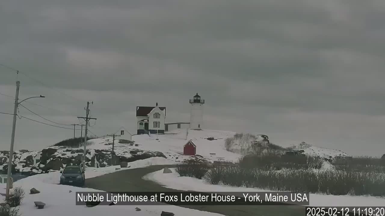 A white lighthouse and adjacent building are situated on a snow-covered hill under an overcast sky.