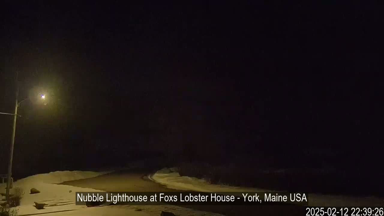 A nighttime scene shows a snow-covered road curving to the right, illuminated by a single streetlight, under a dark sky.