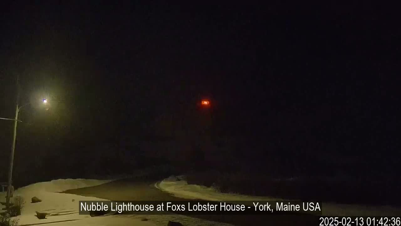 A snow-covered road leads to a distant lighthouse emitting a red light at night.