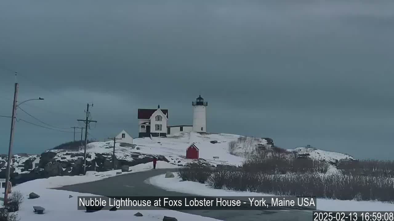 A snow-covered landscape features a lighthouse and accompanying buildings, with a person walking along a road under an overcast sky.