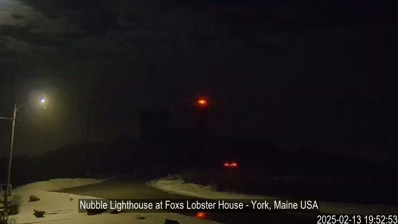A nighttime scene shows a snow-covered shoreline with a lighthouse in the distance under a dark, cloudy sky.