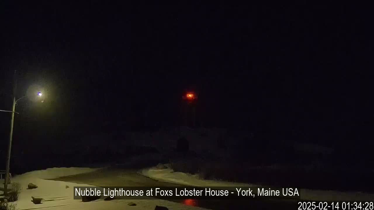 A nighttime view of a lighthouse with a red light, illuminated by a nearby street lamp, in a snowy landscape.
