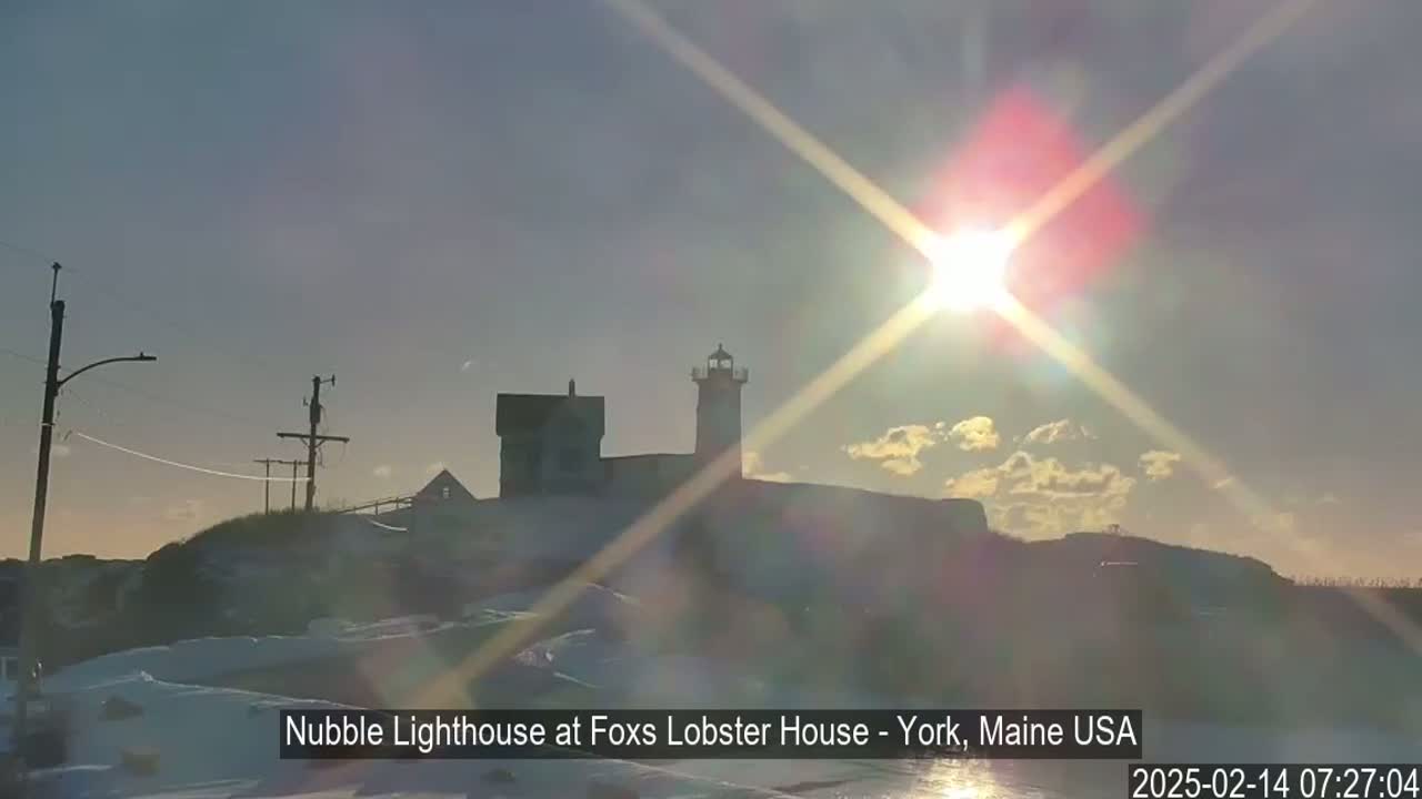 A lighthouse and adjacent buildings are silhouetted against a bright, sun-filled sky, with a snowy landscape in the foreground.