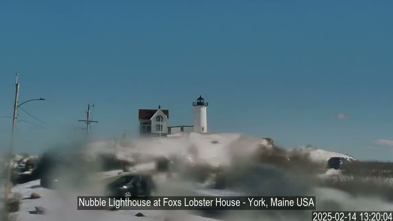 A white lighthouse and a smaller white building are visible in the distance through a snowy, blurry foreground with several vehicles partially obscured by blowing snow under a clear blue sky.