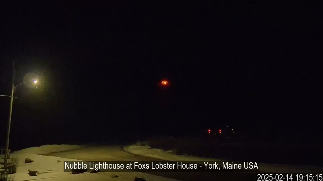 A dimly lit snowy landscape at night features a lighthouse in the distance with its light on.