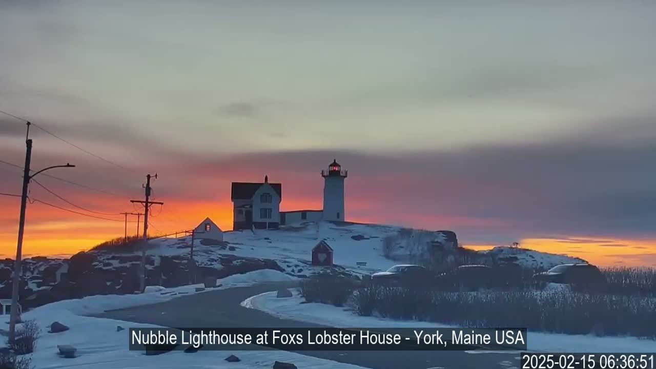 A lighthouse and other buildings sit on a snow-covered hill at sunrise, under a colorful, mostly clear sky.