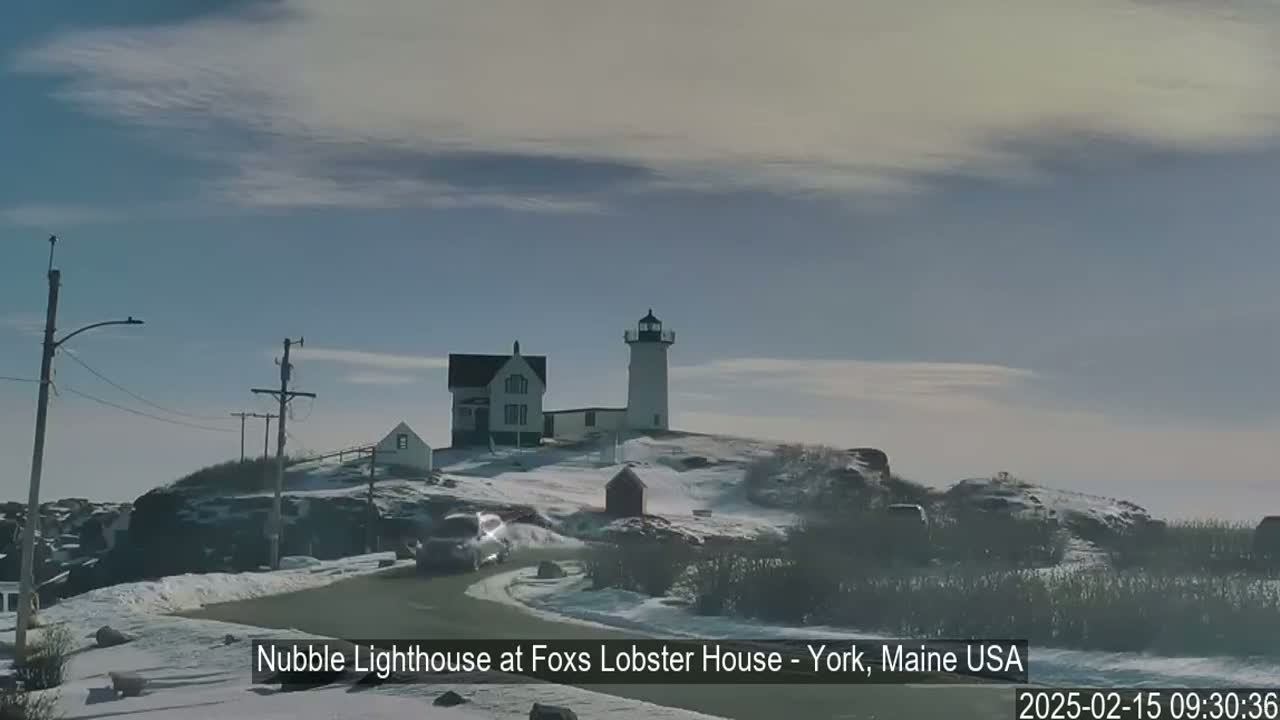 A lighthouse and adjacent buildings sit atop a snow-covered hill under a mostly clear sky.