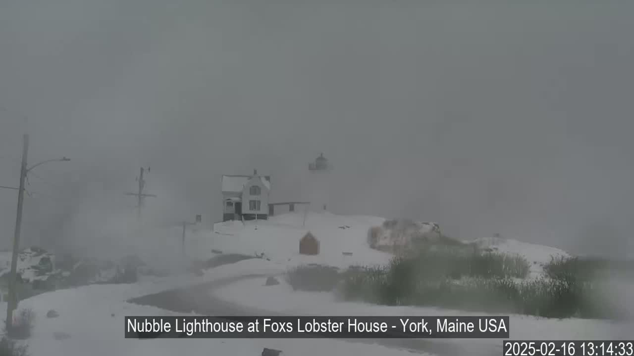 A snow-covered lighthouse and adjacent buildings are barely visible through heavy fog.