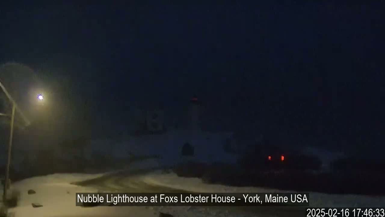 A snow-covered landscape at night shows a lighthouse in the distance under a dark sky.