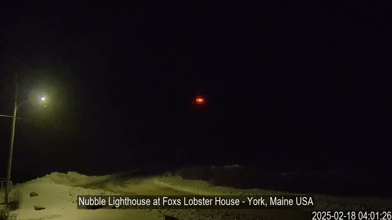 A distant, red light, possibly a lighthouse, is visible in the dark night sky above a snow-covered landscape illuminated by a single streetlight.