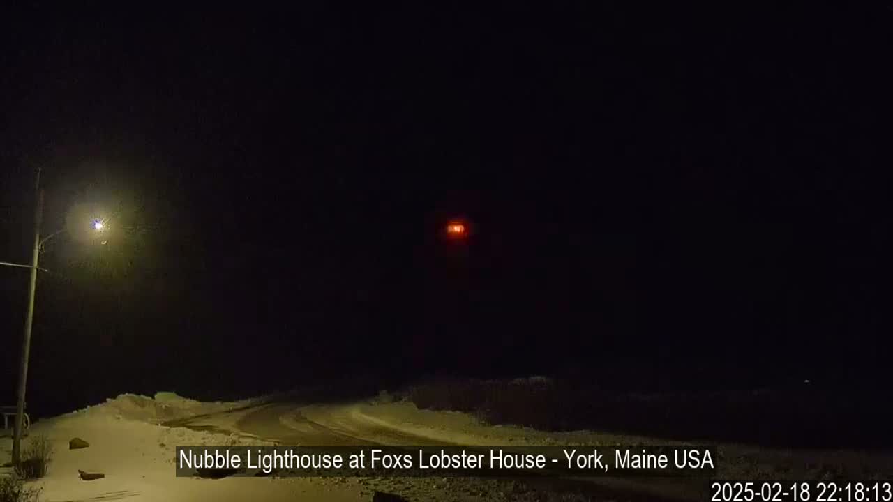 A snow-covered road at night is illuminated by a streetlight, with a distant red light, possibly a lighthouse, visible in the darkness.