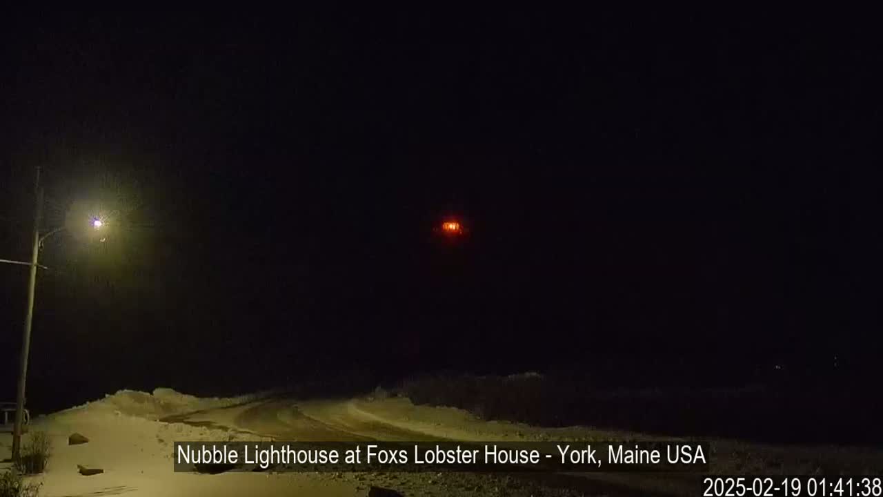 A snow-covered road leads toward a distant, lit lighthouse at night.