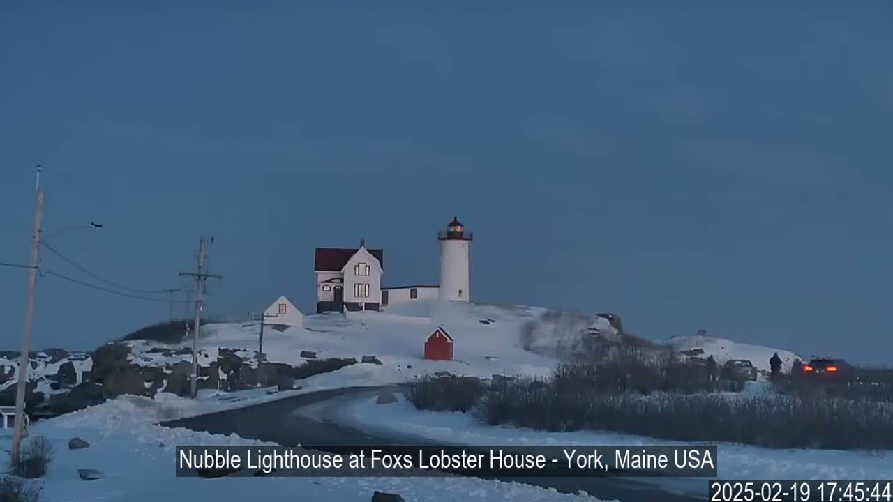 A snow-covered landscape features a lighthouse and accompanying buildings, with a road winding through the scene under a twilight sky.
