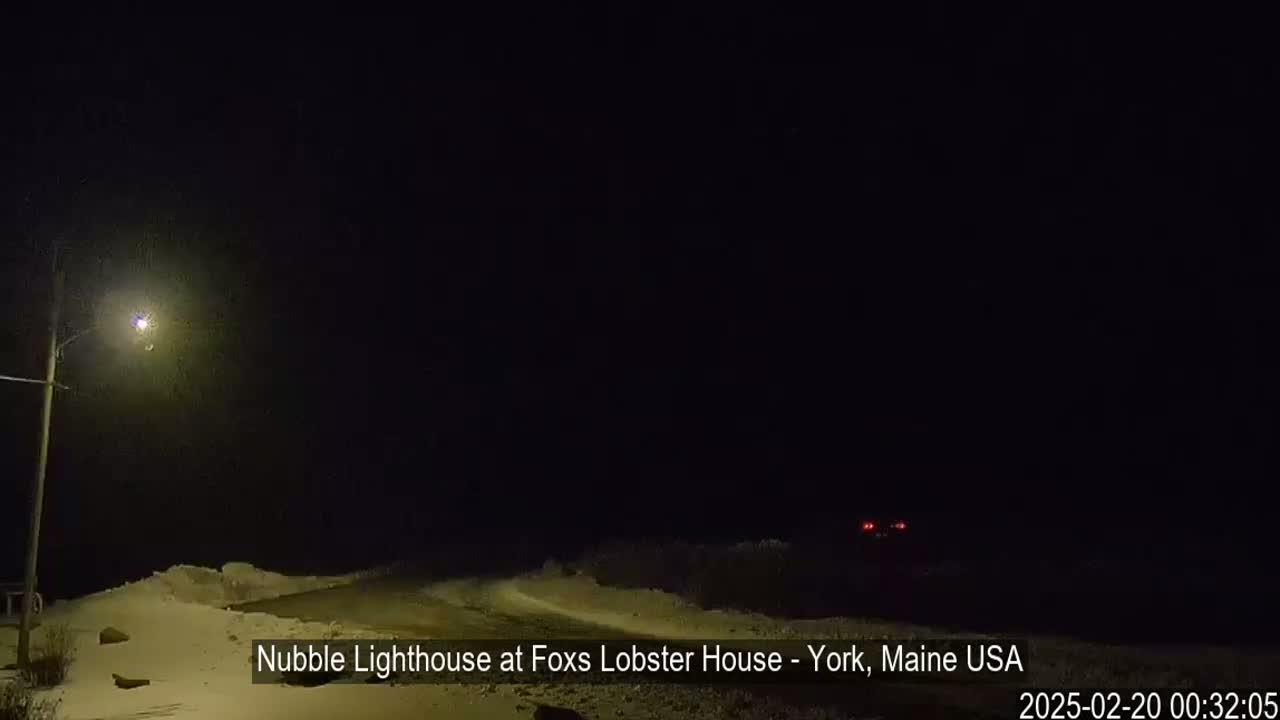 A snow-covered road at night is illuminated by a single streetlight, with a dark sky and a vehicle's taillights visible in the distance.