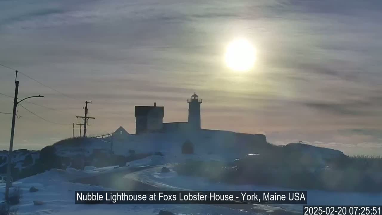 A lighthouse and accompanying buildings stand on a snow-covered hill under a bright, partly cloudy sun.