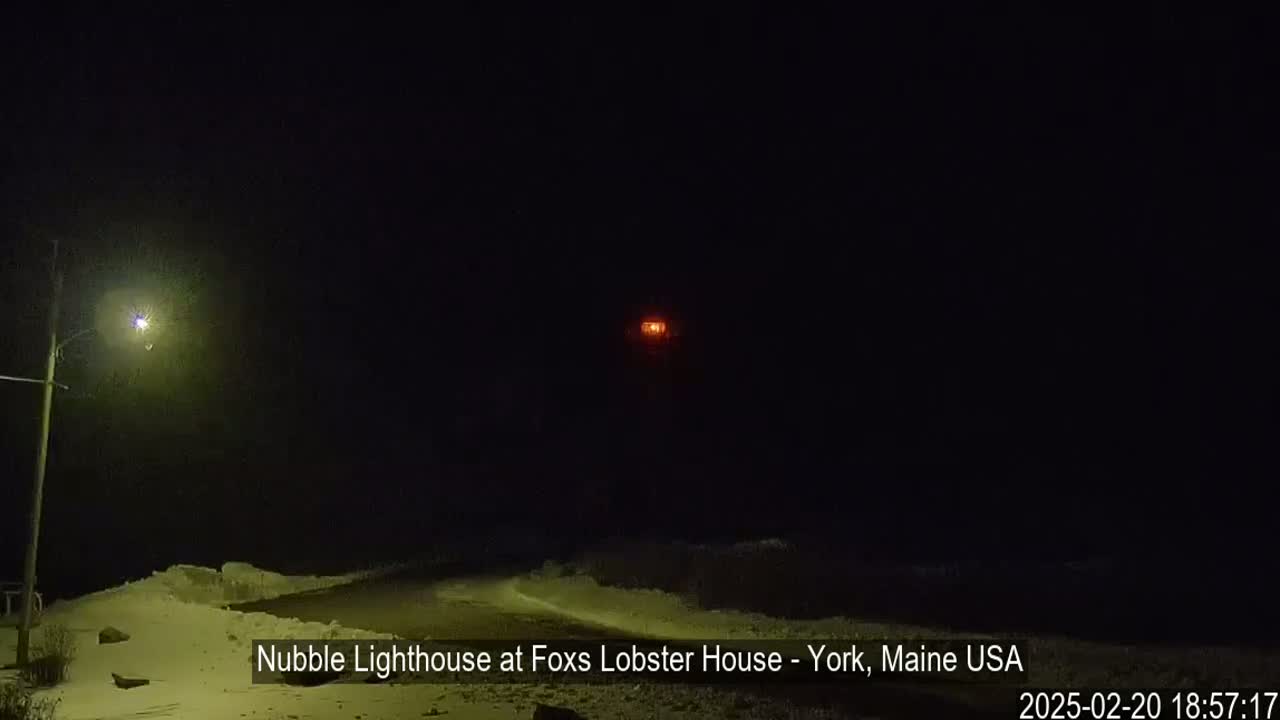 A snow-covered landscape at night is illuminated by a single streetlight and a distant, glowing lighthouse.