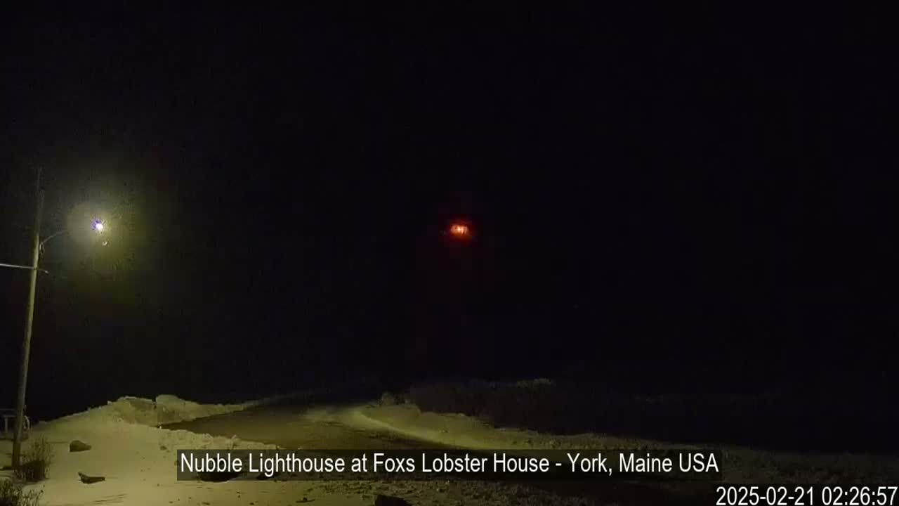 A snow-covered landscape at night is illuminated by a streetlight and a distant lighthouse.