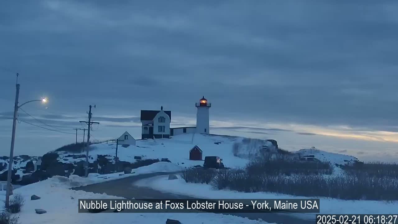 A snow-covered landscape features a lighthouse with a lit lantern, a smaller building nearby, and a vehicle parked on a road.