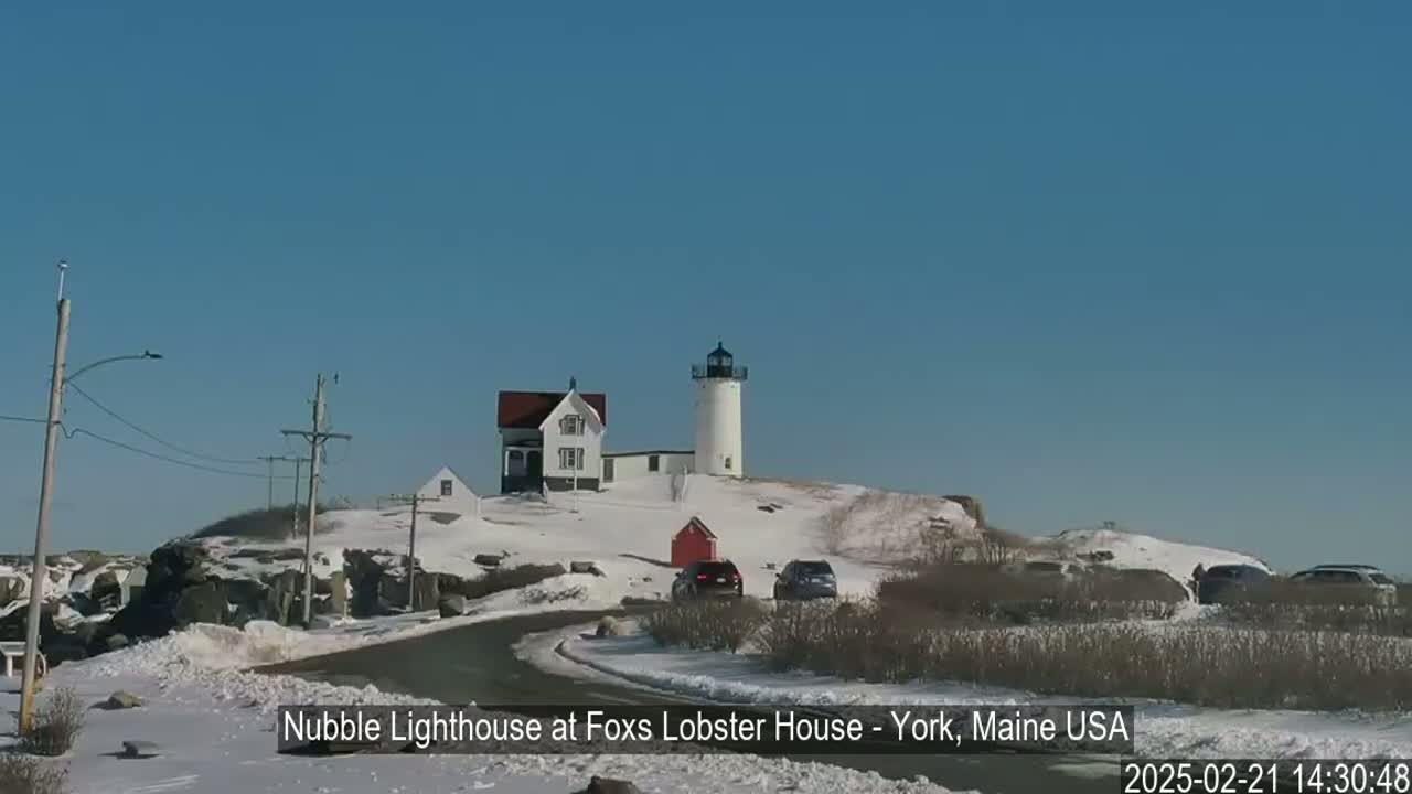 A lighthouse and keeper's house sit atop a snow-covered hill, with cars parked nearby on a partly icy road under a clear blue sky.