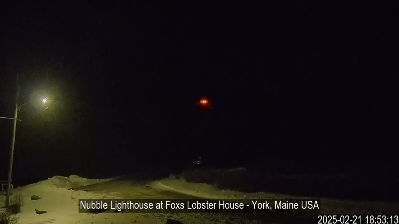 A nighttime view of a snow-covered landscape shows a distant, lit lighthouse and a streetlight in the foreground under a dark, clear sky.