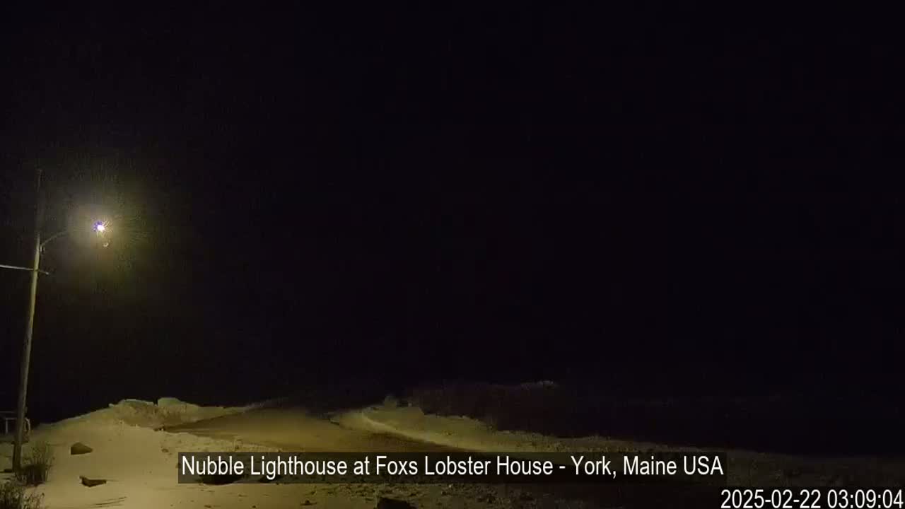 A nighttime scene shows a snow-covered shoreline illuminated by a single lamppost under a dark sky.
