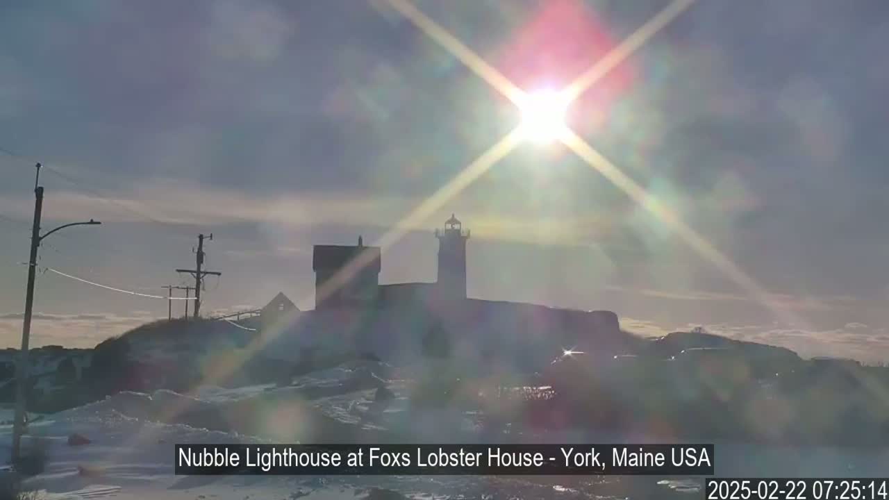 A snow-covered landscape features a lighthouse and other buildings silhouetted against a bright, sun-filled sky.