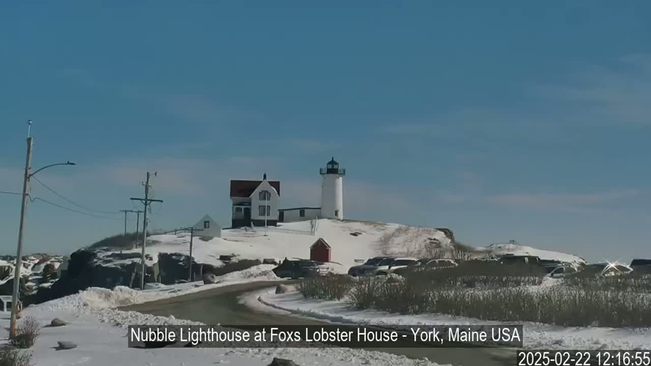 A white lighthouse and accompanying building are situated on a snow-covered hill overlooking a road with parked cars under a clear blue sky.