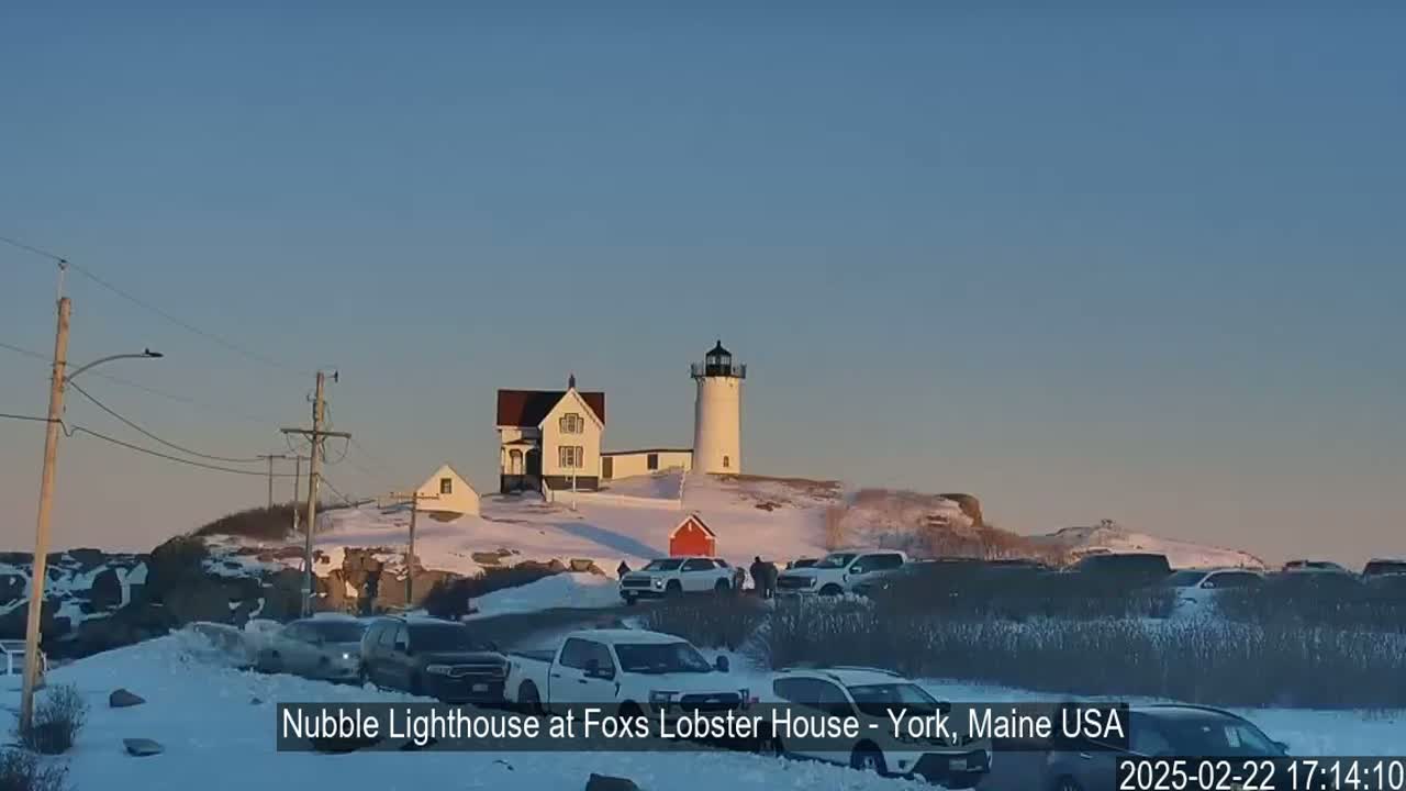 A lighthouse and accompanying buildings sit atop a snow-covered hill with several cars parked nearby, under a clear, twilight sky.
