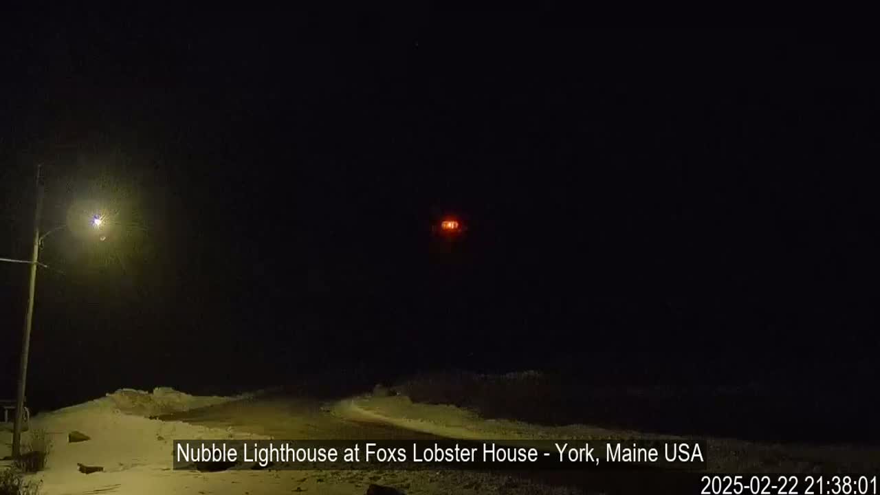 A snow-covered, nighttime scene shows a lighthouse's red light in the distance, illuminated by a streetlamp in the foreground.