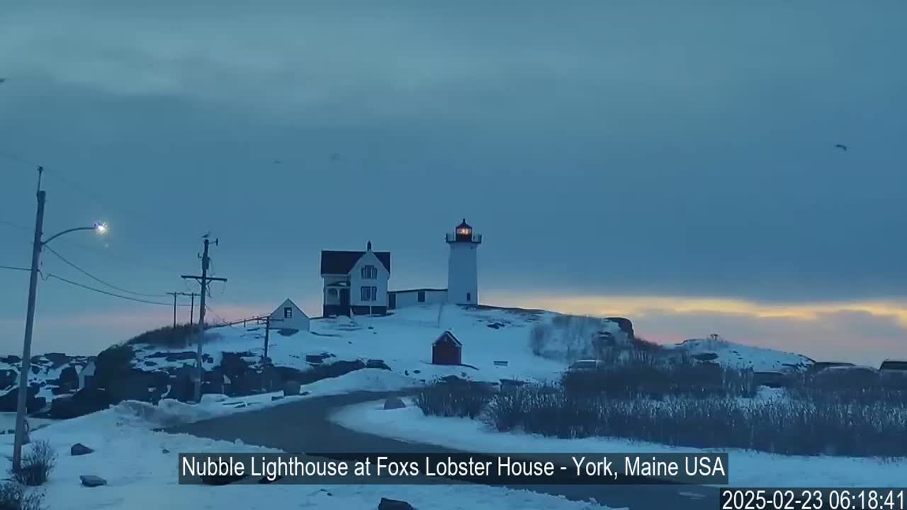 A snow-covered lighthouse and adjacent buildings sit atop a rocky hill at dawn under a clear, dusky sky.
