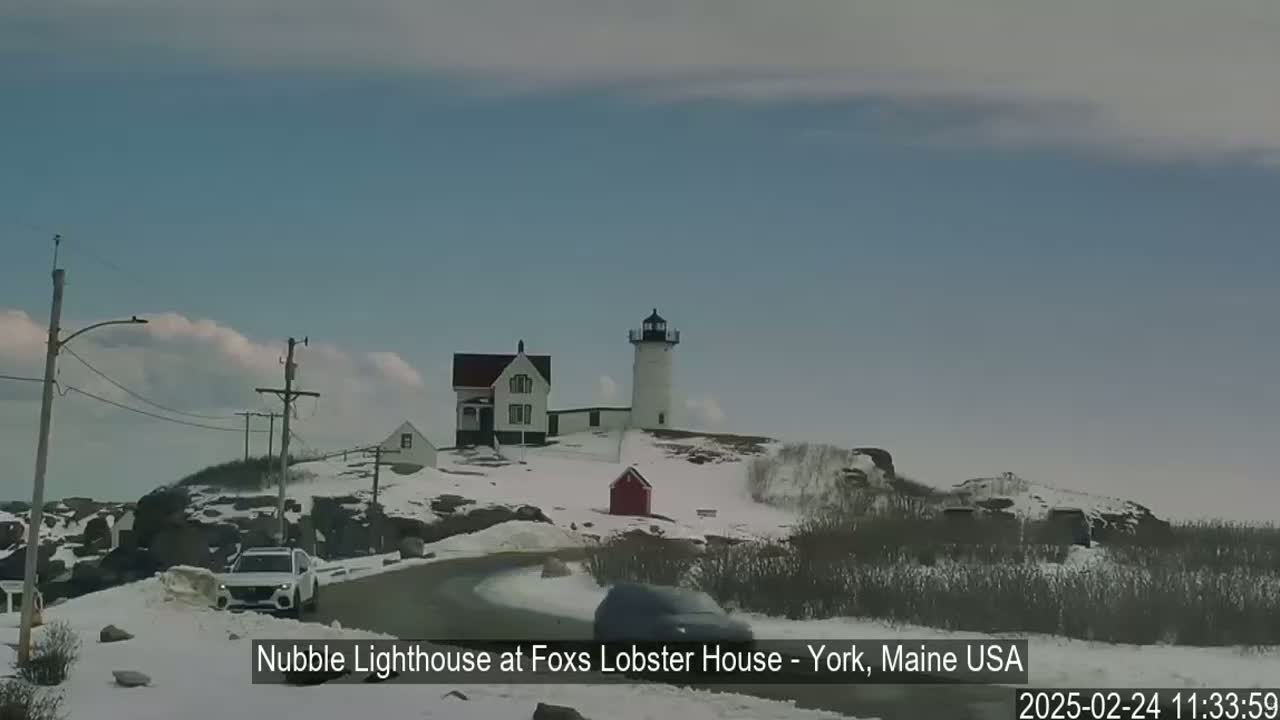 A lighthouse and houses are situated on a snow-covered rocky outcrop under a mostly clear sky.