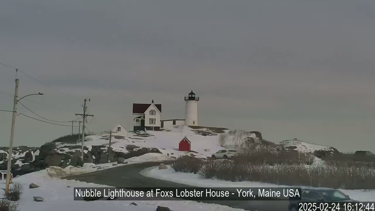 A white lighthouse stands on a snow-covered rocky outcrop, with a white house and a small red building nearby under an overcast sky.