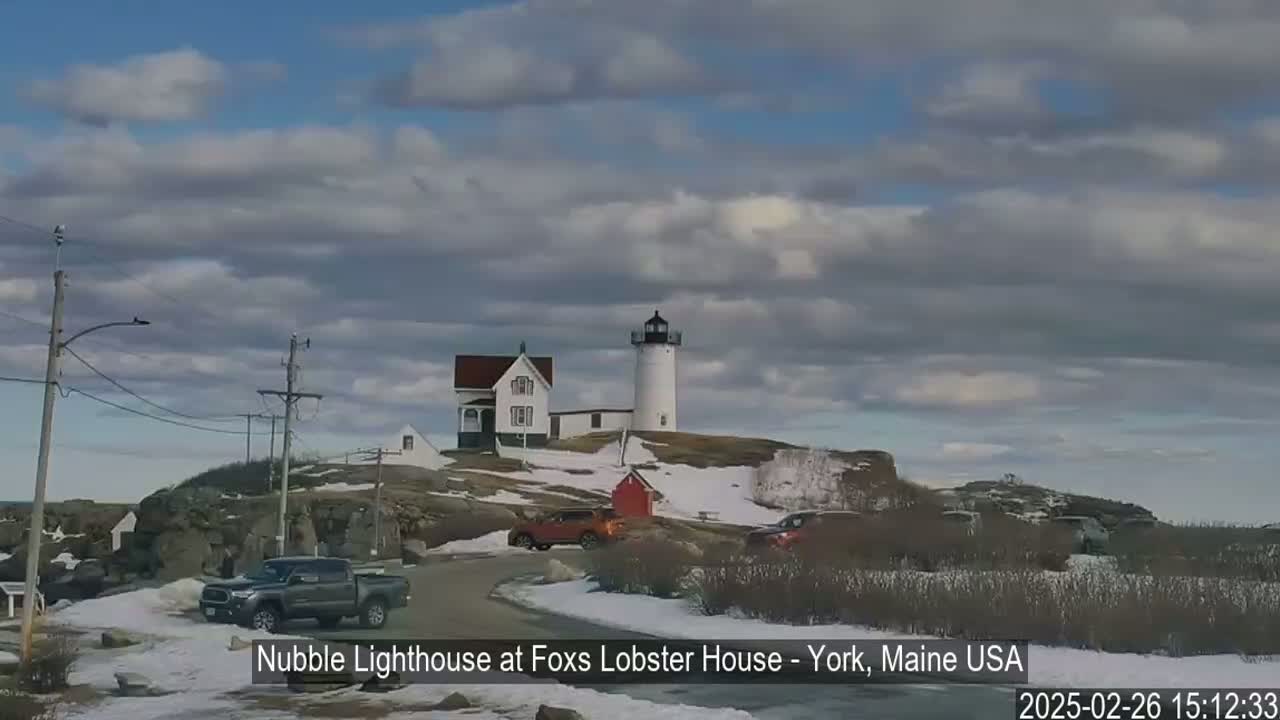 A lighthouse and keeper's house sit atop a snow-dusted rocky hill under a partly cloudy sky.