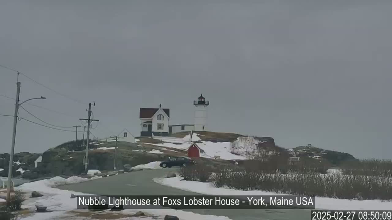 A lighthouse and keeper's house sit on a snow-covered, rocky outcropping under an overcast sky.