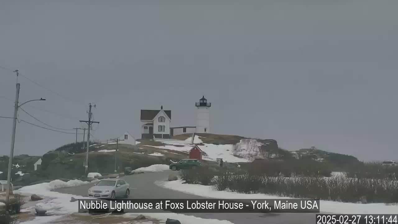 A lighthouse and a house sit on a snow-dusted hilltop under an overcast sky, with cars parked nearby on a road.