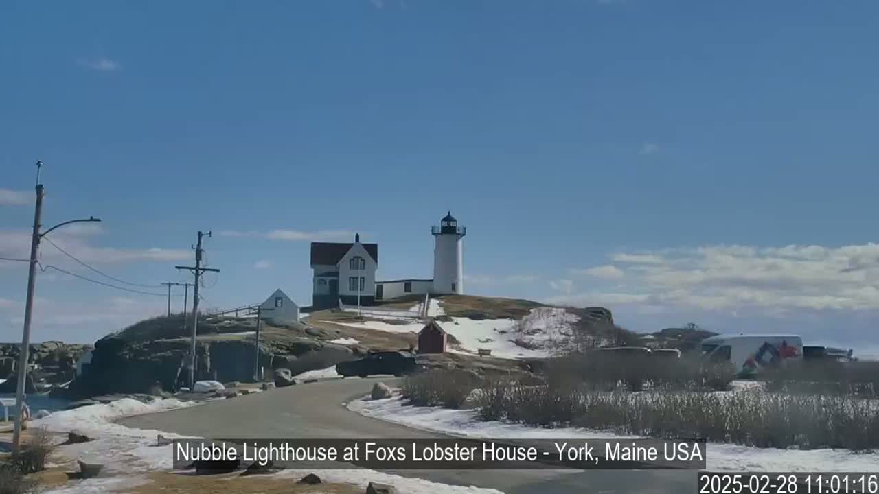 A lighthouse and adjacent buildings sit on a rocky, snow-dusted coastline under a mostly clear blue sky.