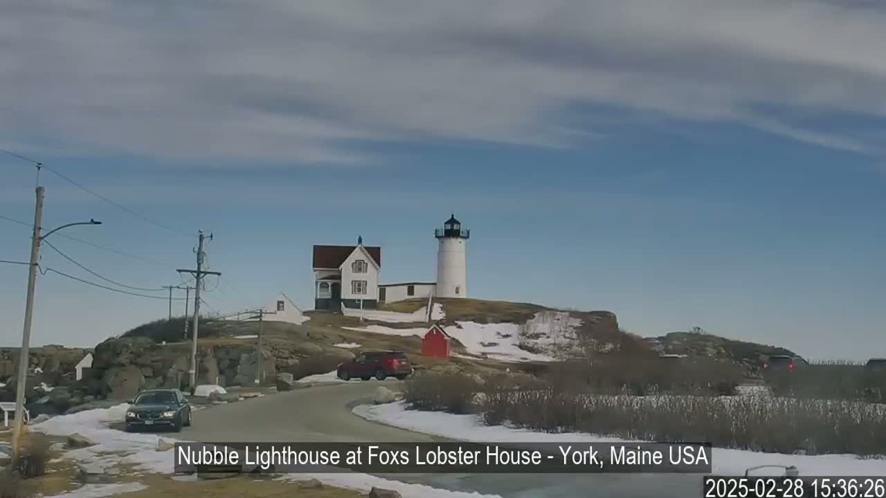 A white lighthouse and a white house sit atop a rocky hill with patches of snow, under a mostly clear blue sky.