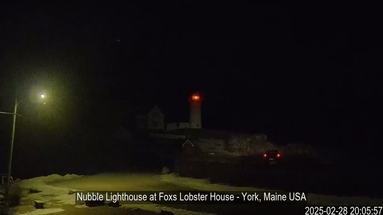 A lighthouse with a lit lamp shines brightly at night in a snowy landscape illuminated by a streetlight.