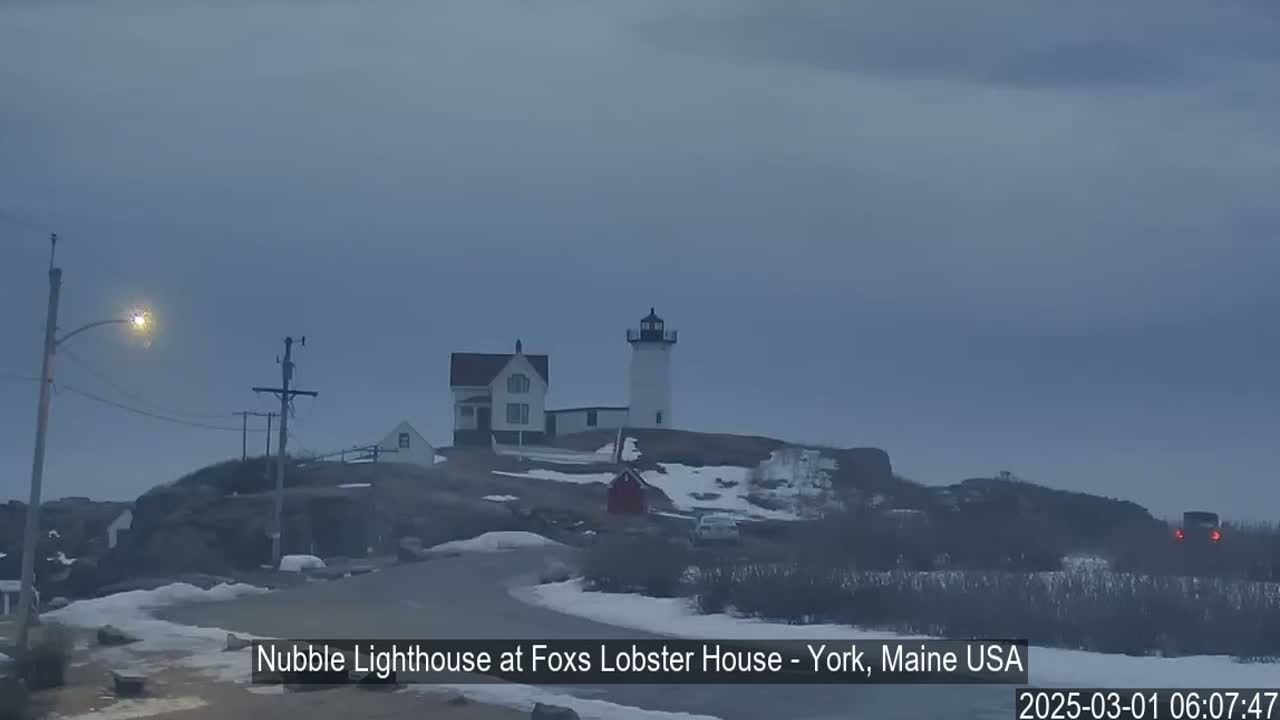 A lighthouse and a smaller building sit on a snow-dusted hilltop under a cloudy, gray sky.