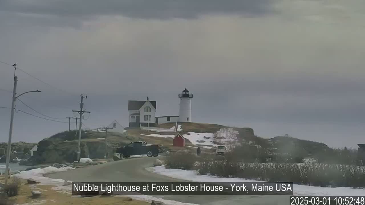 A lighthouse and other buildings sit atop a snow-dusted rocky outcrop on an overcast day.