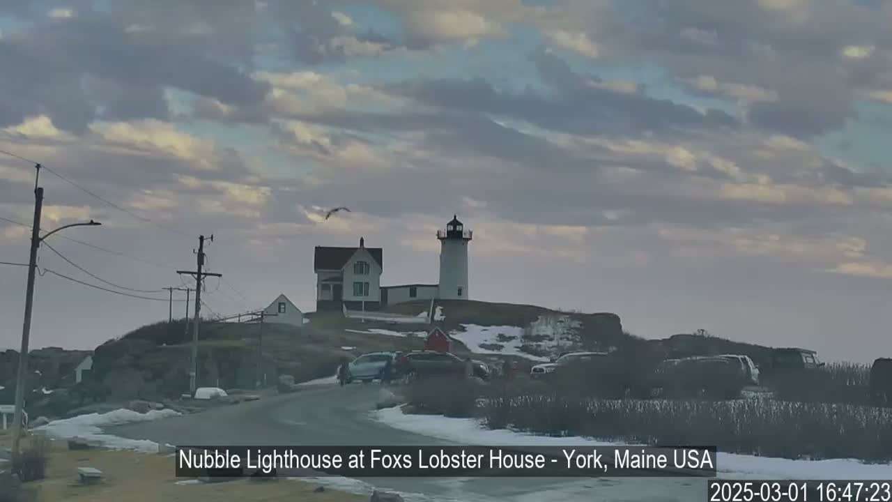 A lighthouse and adjacent house sit atop a snow-dusted hill under a cloudy sky.