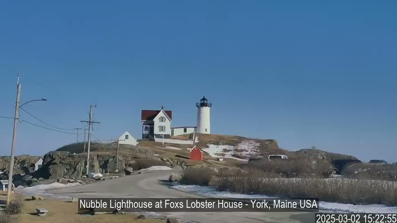 A lighthouse and keeper's house sit atop a rocky hill under a clear blue sky, with patches of snow and ice visible on the ground.