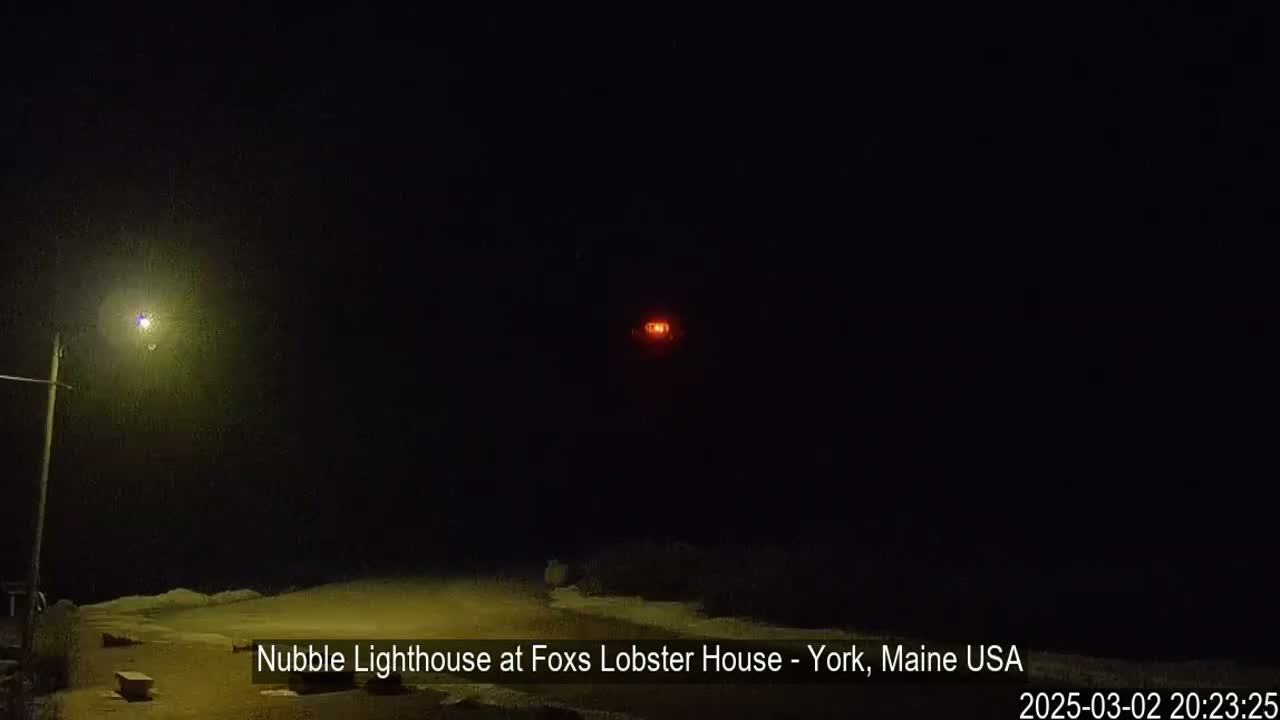 A nighttime view of a distant, lit lighthouse and a nearby streetlight, with snow on the ground.