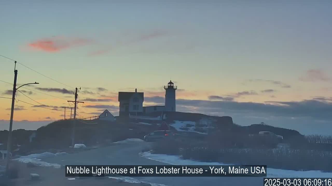 A lighthouse and adjacent buildings are situated on a hill overlooking a road at sunrise, with a pinkish hue in the sky and patches of snow on the ground.
