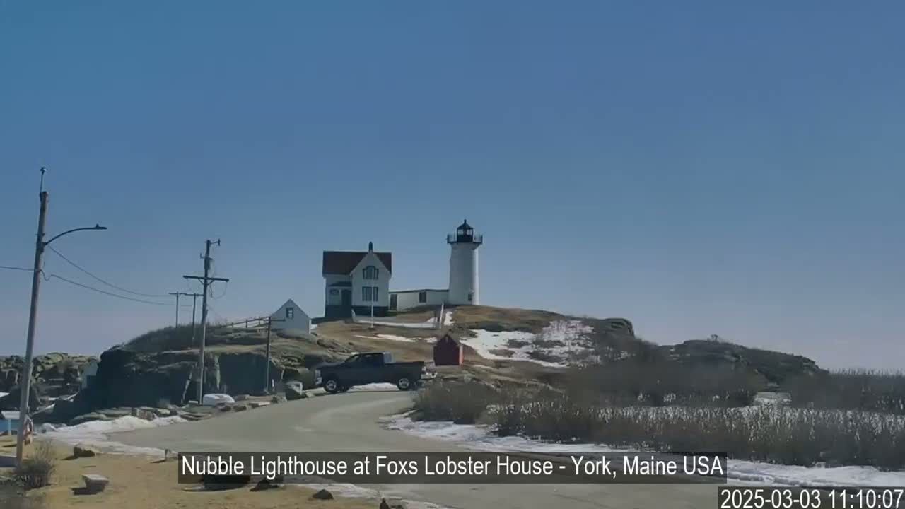 A white lighthouse and keeper's house sit atop a rocky hill under a clear blue sky, with patches of snow visible on the ground.