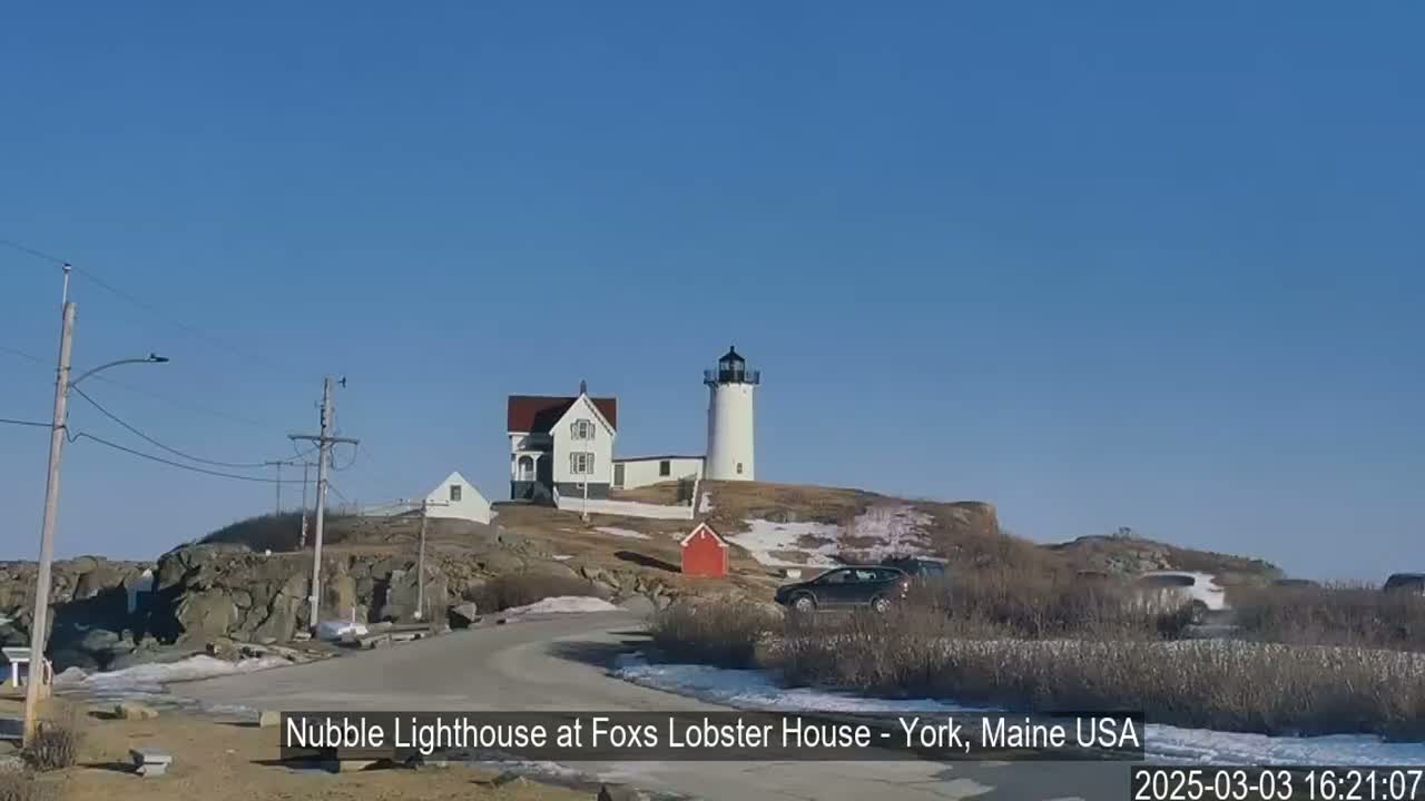 A white lighthouse and a house sit atop a rocky hill under a clear blue sky, with patches of snow on the ground and a few cars parked nearby.