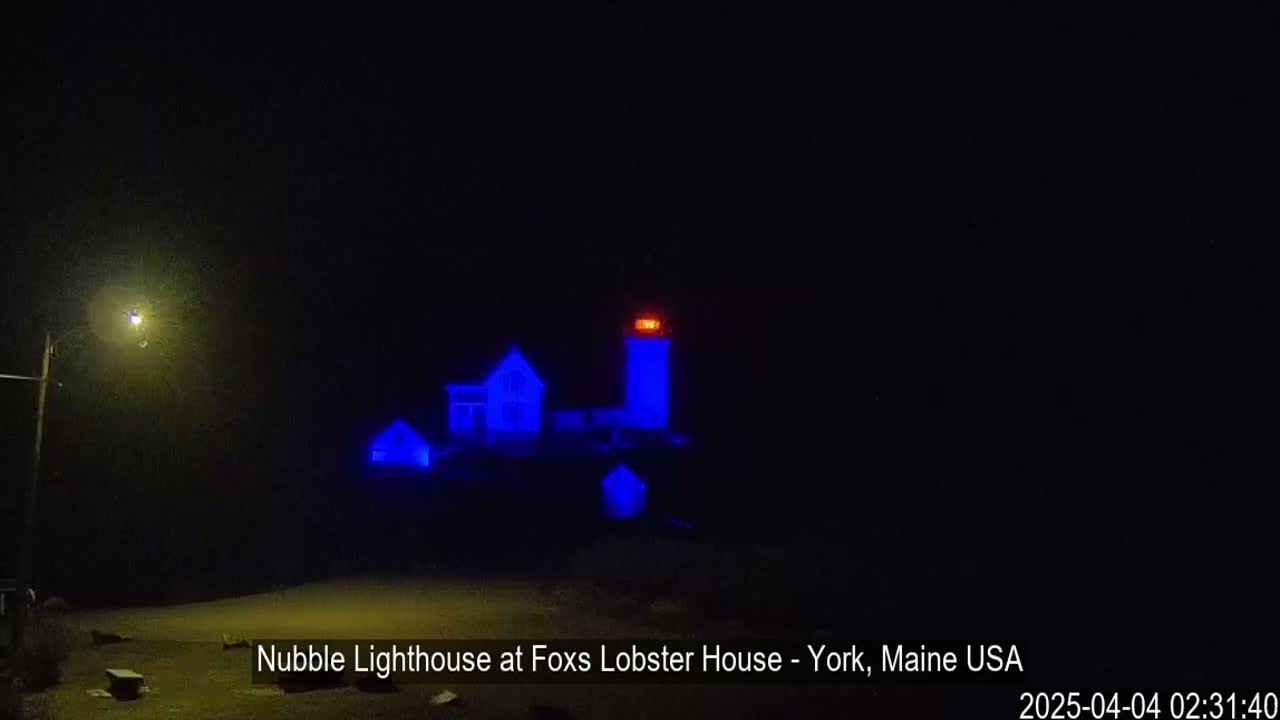 At night, a lighthouse and nearby buildings are illuminated in blue light.