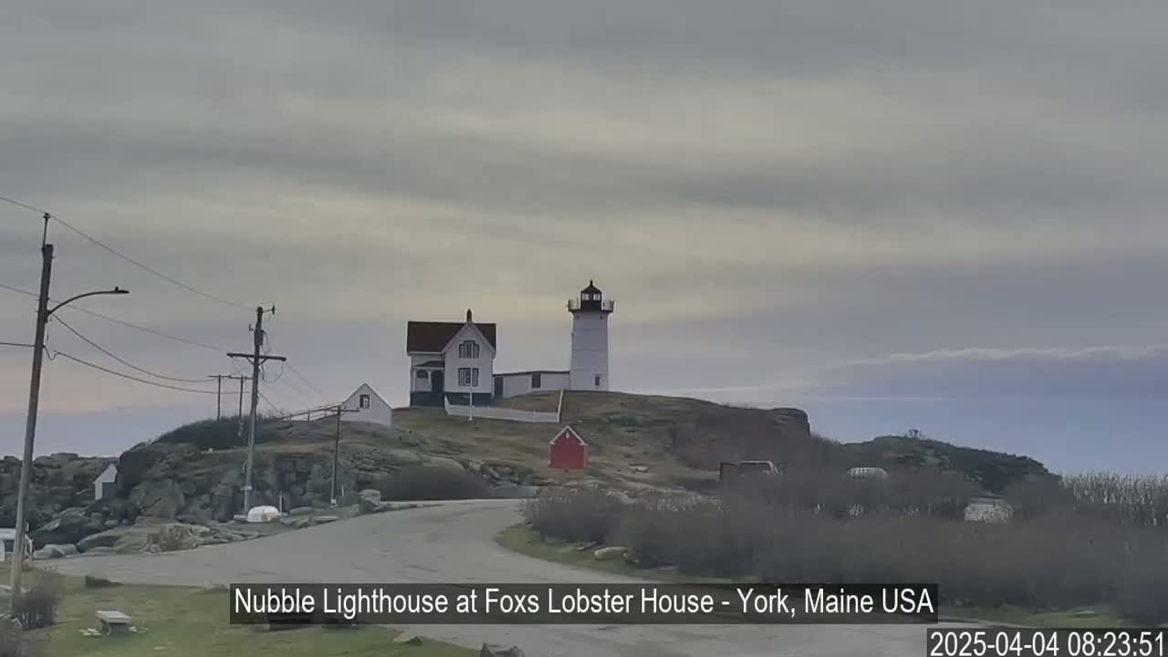 A lighthouse and a house sit on a rocky hill overlooking a road under an overcast sky.