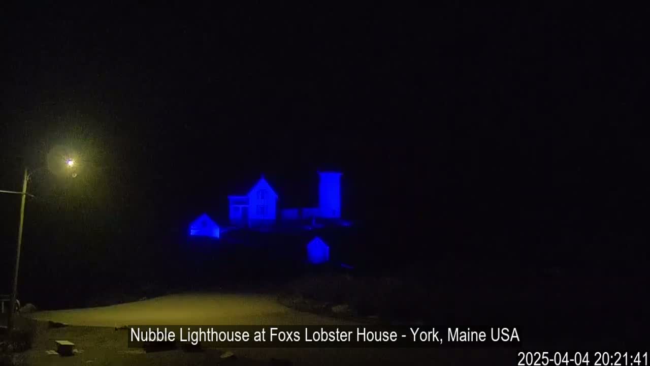 A lighthouse and adjacent buildings are illuminated in blue at night.