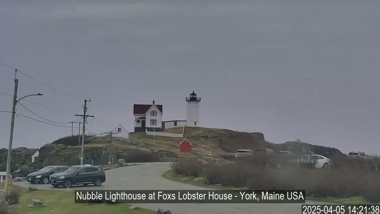 A white lighthouse and a smaller white house sit atop a rocky hill under an overcast sky, with several cars parked nearby and a road curving past.